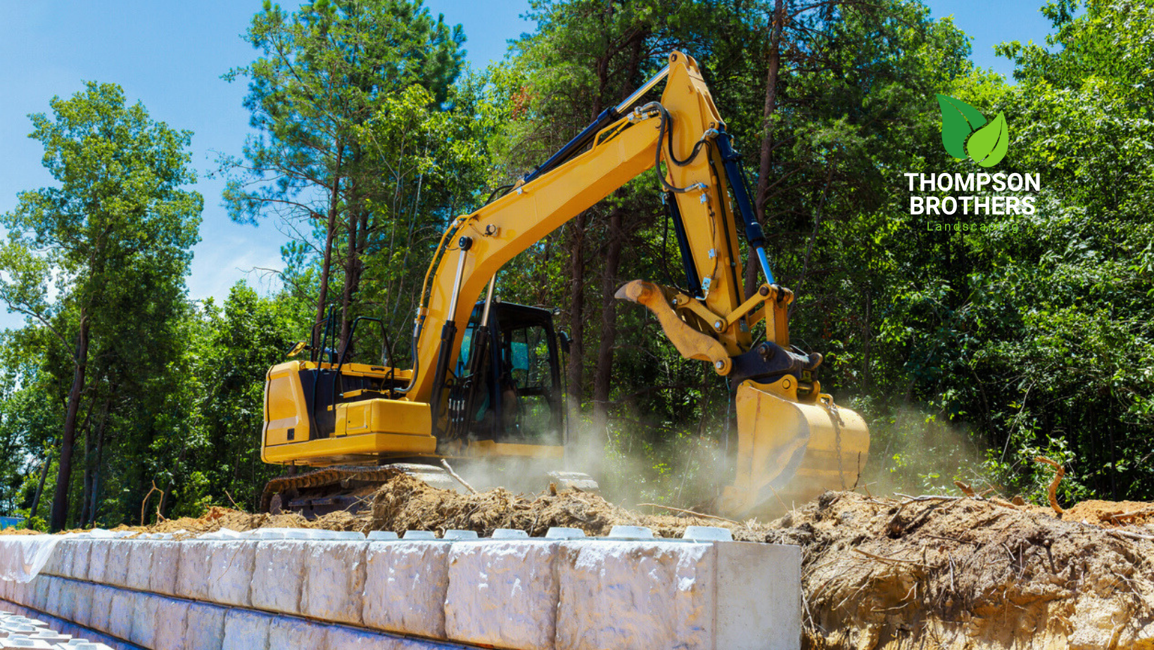A yellow excavator digging dirt behind a newly constructed white stone retaining wall, with green trees in the background.