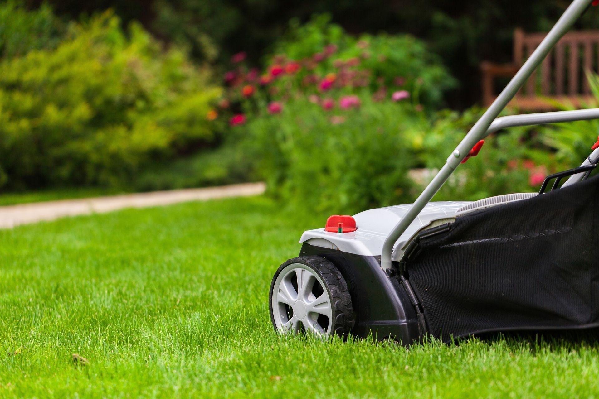 A side view of a lawnmower on a vibrant green lawn with a garden and flower bed in the blurred background.