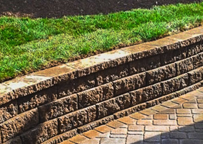 A stone retaining wall separates a grassy upper lawn from a patterned, light-brown paver patio.