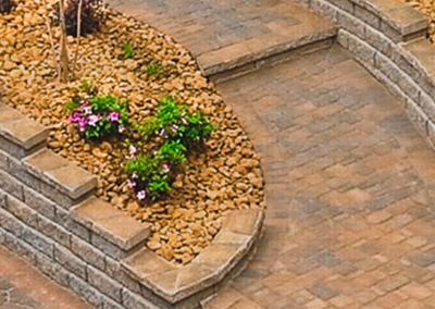 A curved stone retaining wall borders a landscape bed with gravel and flowers, next to a paved stone walkway.