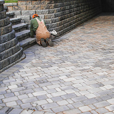 A construction worker in tan overalls kneels to install stone pavers near a curved retaining wall.