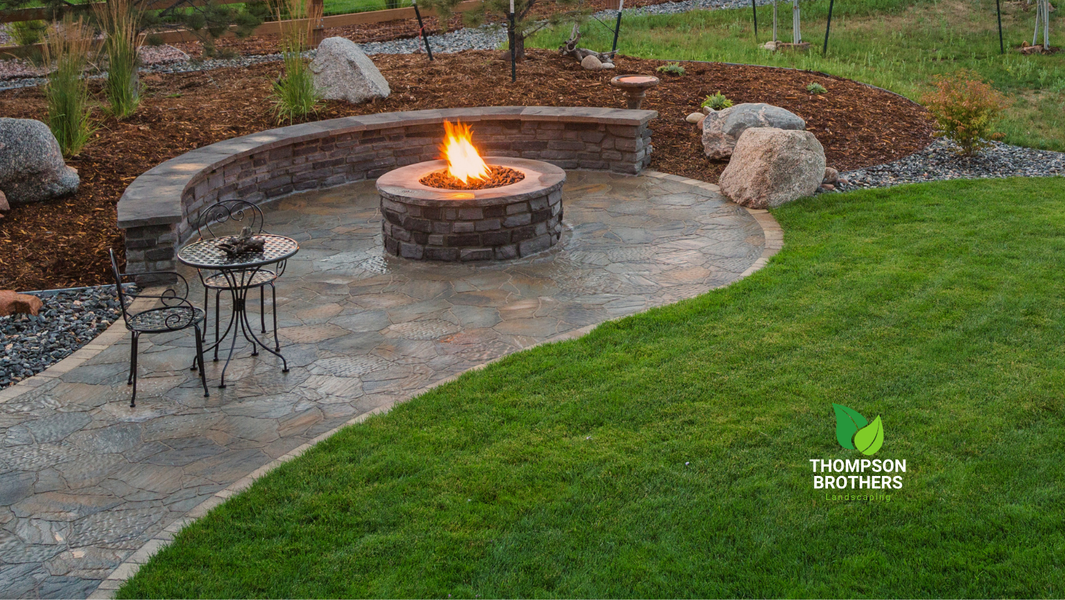A backyard stone patio featuring a circular fire pit with a lit flame, adjacent curved seating, and a nearby metal table.
