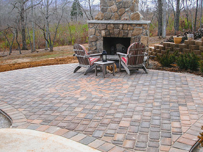 Two Adirondack chairs sit on a paver patio in front of a stone outdoor fireplace in a wooded yard.