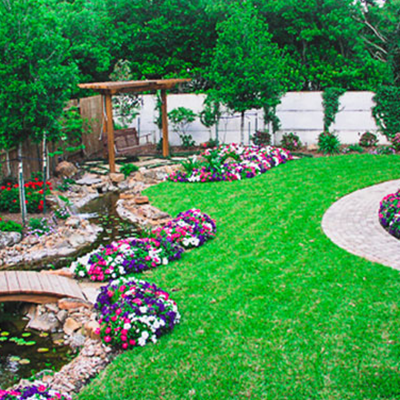 A lush garden with a curved stone path, a small wooden bridge over a stream, flower beds, and a swing under a pergola.