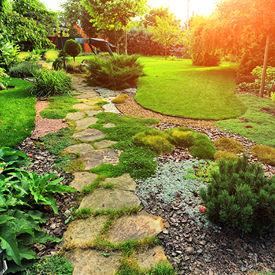 A stone path winds through a lush, sunny garden with green lawn, shrubs, and mulch beds under warm, glowing sunlight.