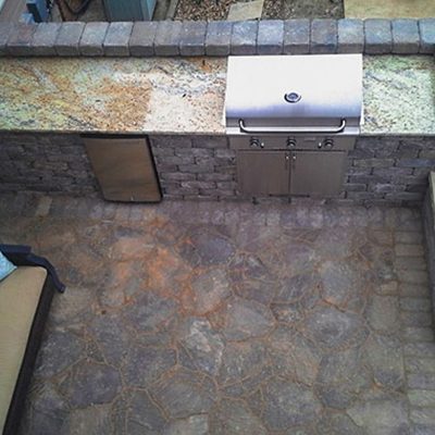 An outdoor kitchen with a stainless steel grill, stone counter, and brick base on a patio paved with irregular flagstone.