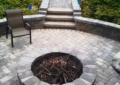 A stone patio featuring a circular fire pit filled with twigs, a brown lawn chair, and a short staircase leading upward.