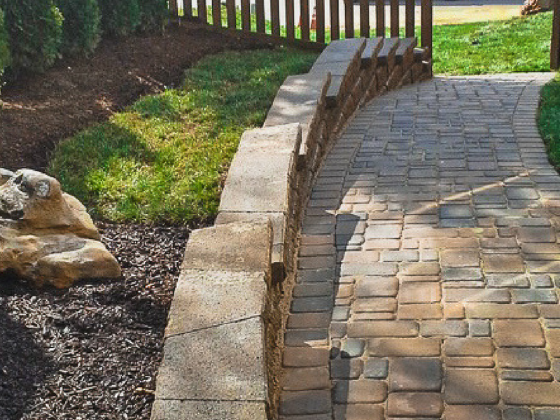 A stone-paved walkway bordered by a stacked block retaining wall, set beside a mulched garden bed and wooden fence.