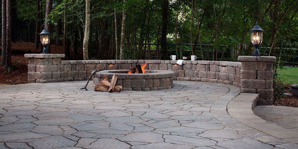 Outdoor fire pit area with stone seating, pavers, and pillar lights set in a wooded backyard.