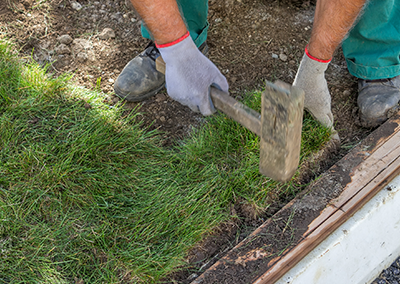 A person in work gloves uses a wooden mallet to flatten a piece of new sod next to a wooden garden border.