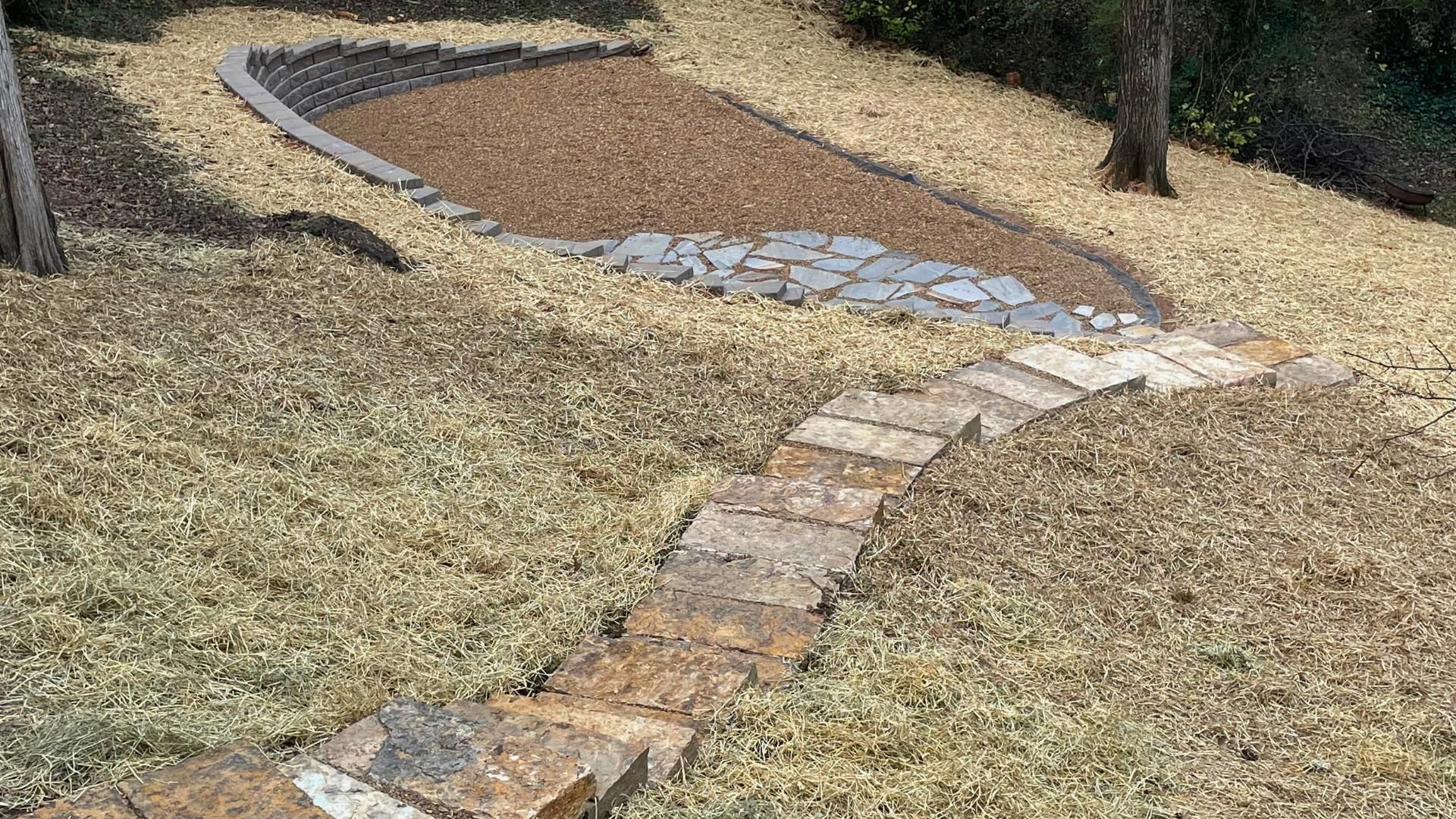 A stone pathway leads up a grassy slope toward a curved stone retaining wall surrounding a gravel garden bed.