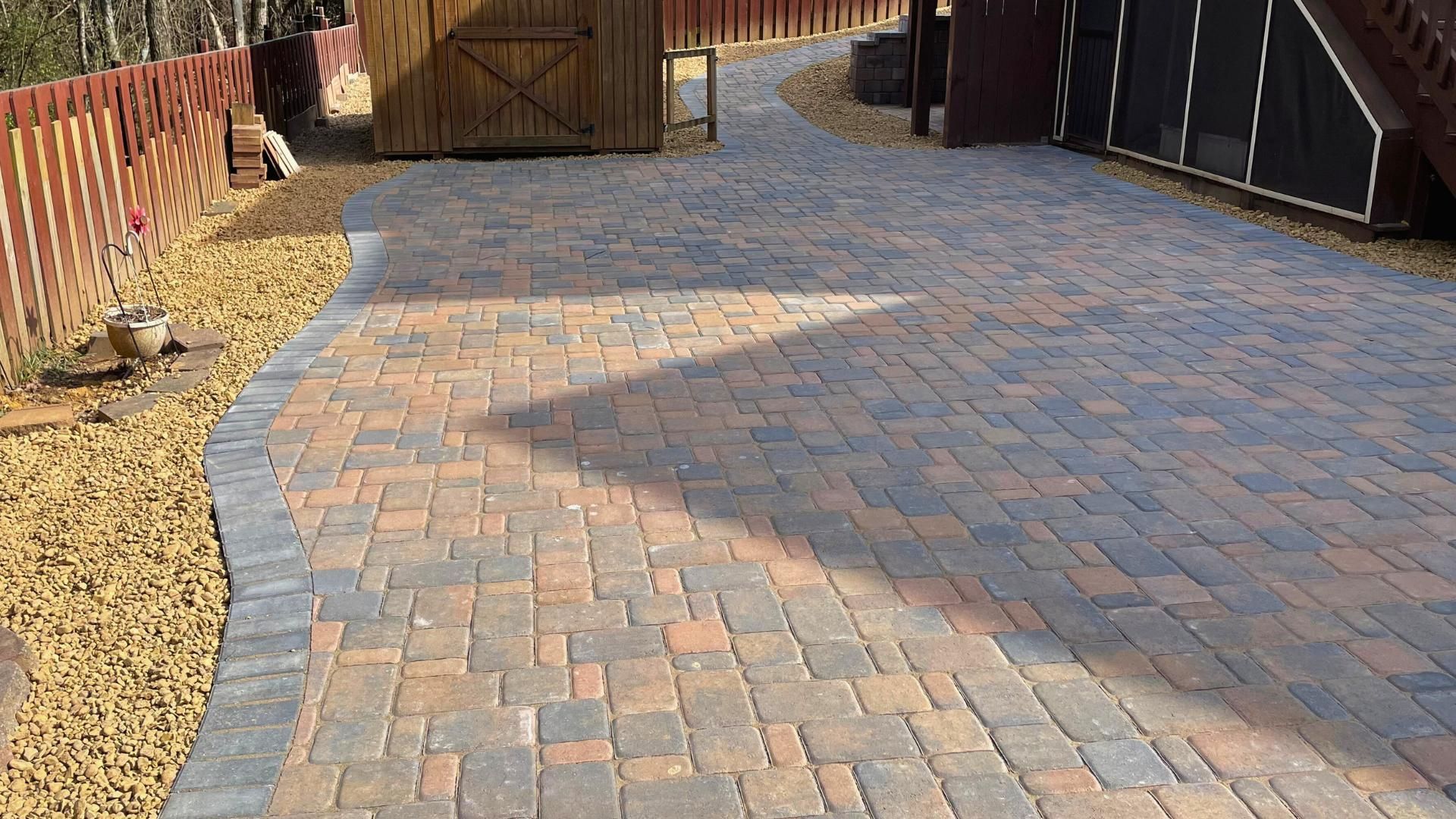 A multi-colored paver patio with a dark border, bordered by a wooden fence, gravel, and a storage shed.