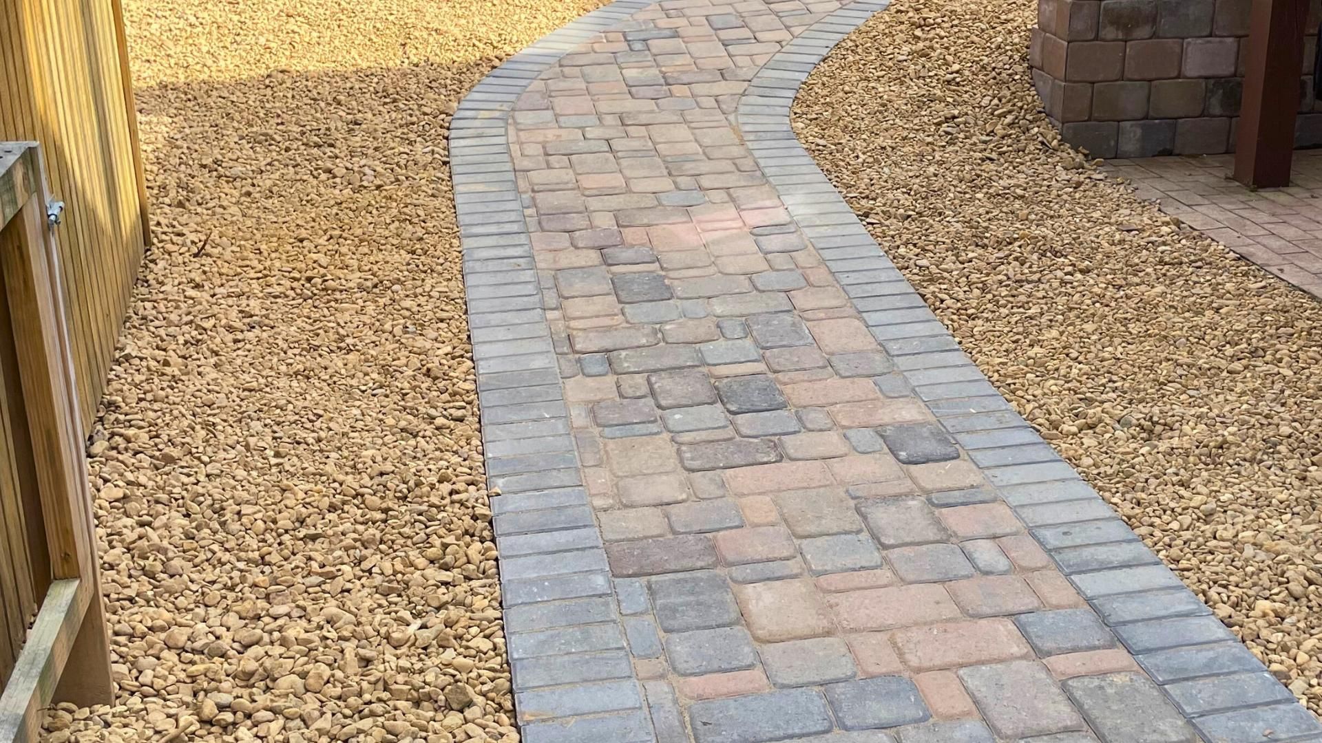 A winding walkway made of multicolored rectangular pavers, bordered by gray bricks and surrounded by light brown gravel.