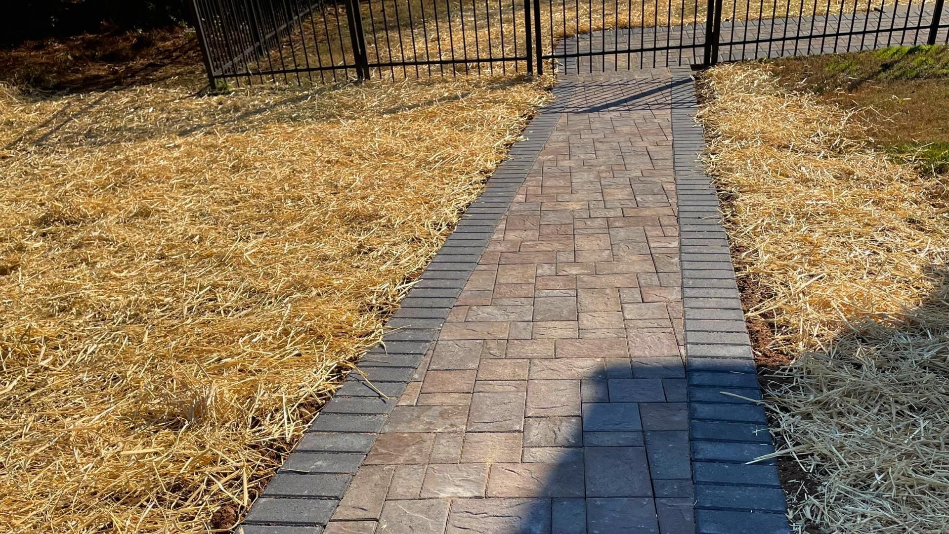 A brick pathway with a dark border runs through a yard with light-colored ground cover and a metal fence in the background.