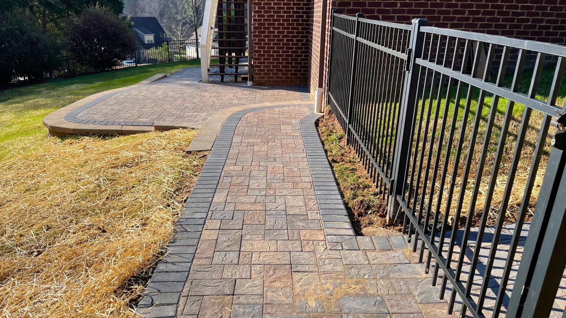 A paved stone walkway with dark border bricks leads to a matching patio next to a brick building and black metal fence.