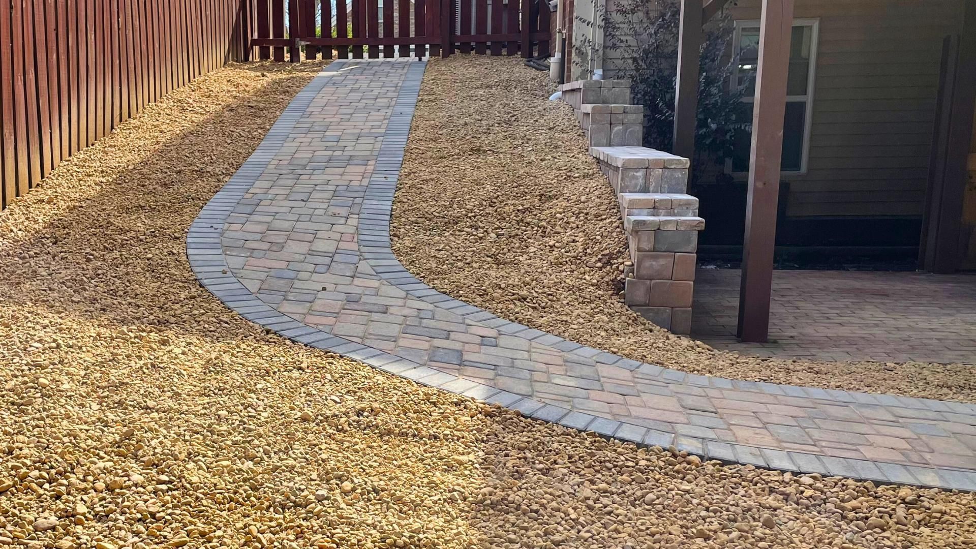 A curved stone paver walkway winds through a yard covered in light brown gravel, leading toward a wooden patio structure.