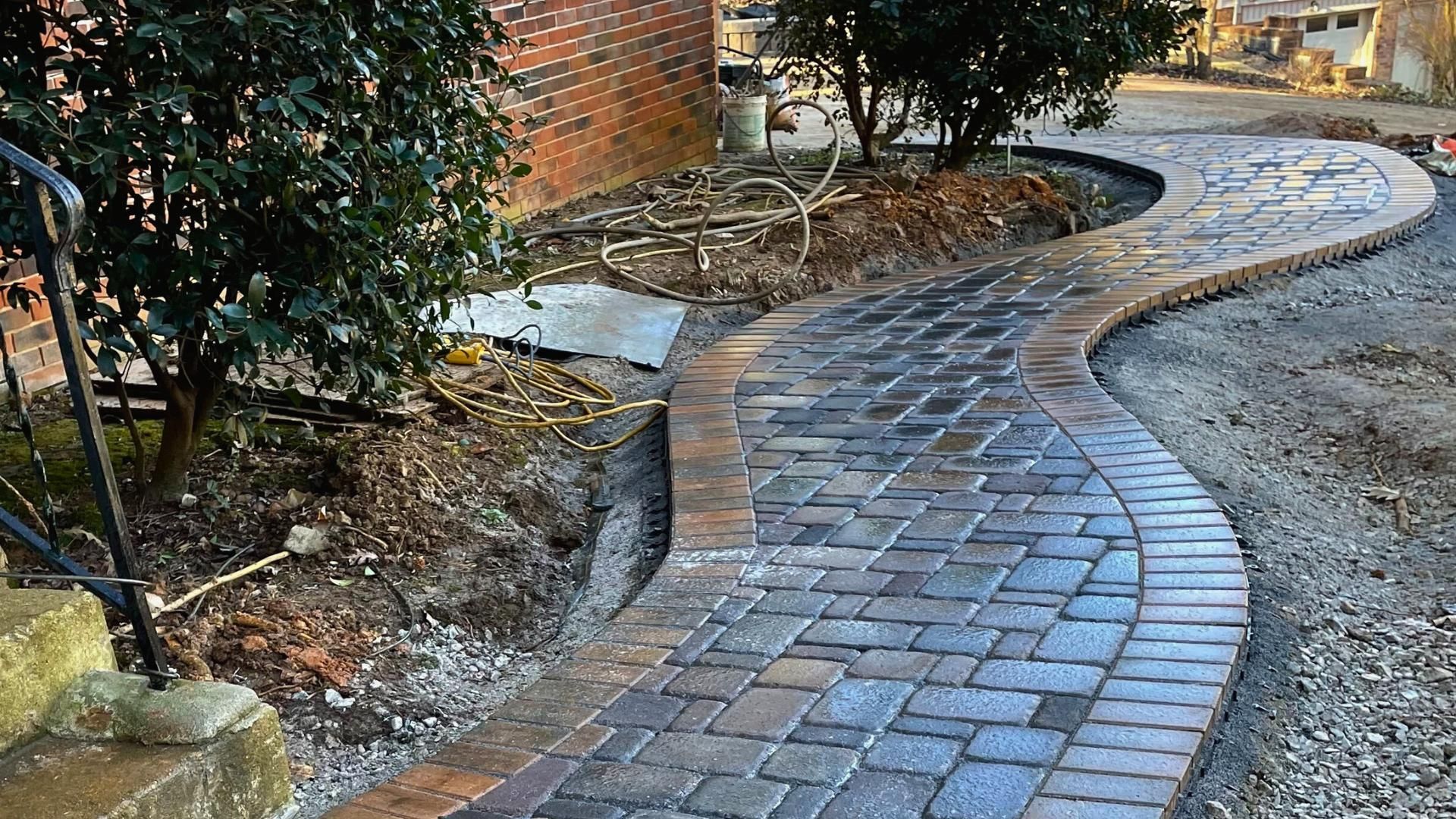 A curved outdoor walkway made of multi-colored rectangular pavers with a matching border, situated next to shrubs and trees.