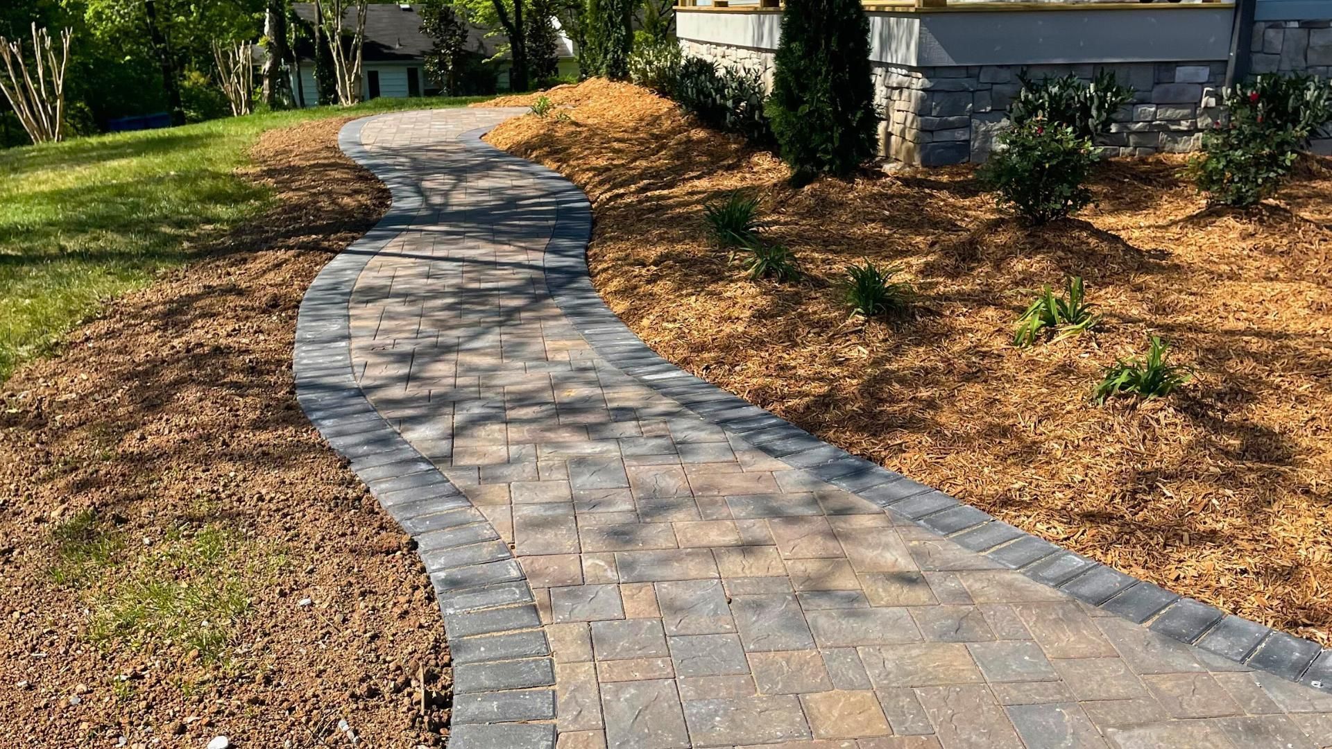 A curved stone paver walkway bordered by dark bricks, flanked by mulched garden beds and a grassy lawn.