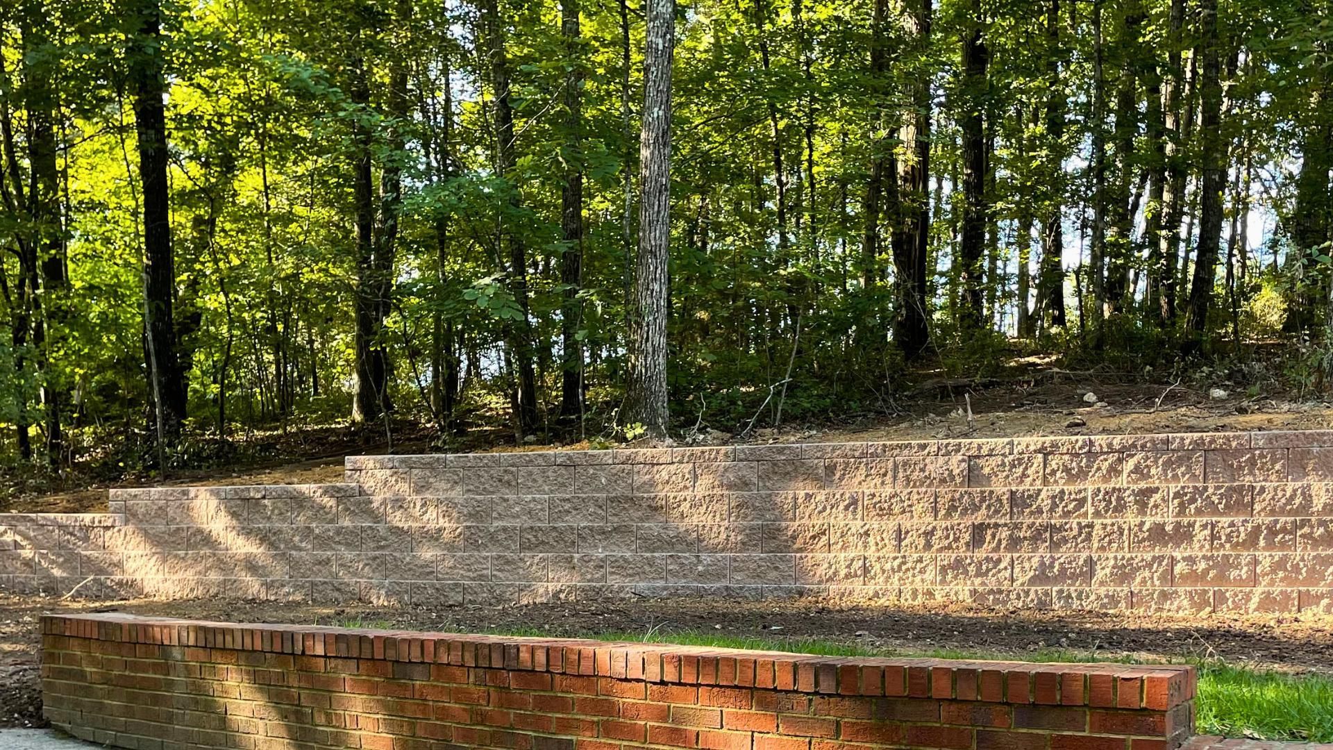 A tiered stone seating area and a low brick wall set against a backdrop of dense, sunlit green trees.