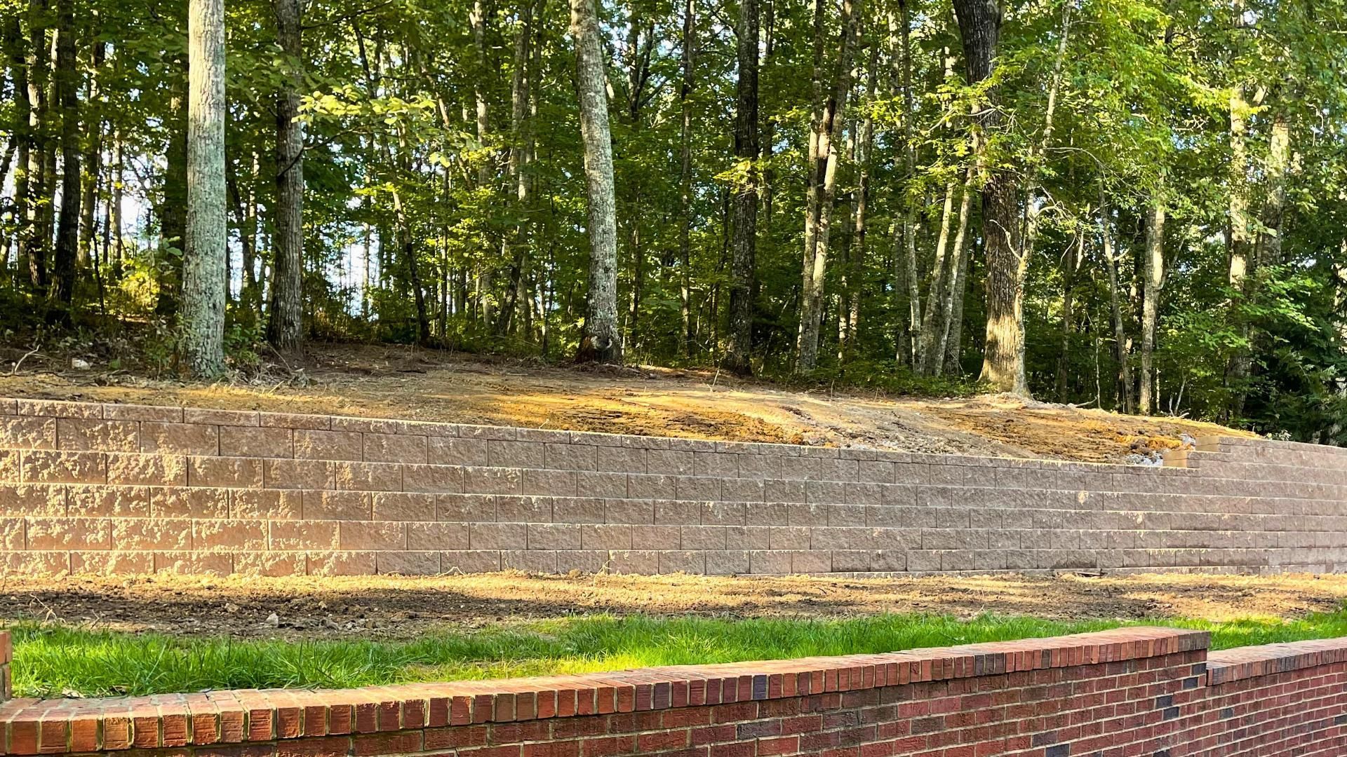 A tiered retaining wall made of reddish-brown blocks in front of a wooded area with trees and scattered dry leaves.