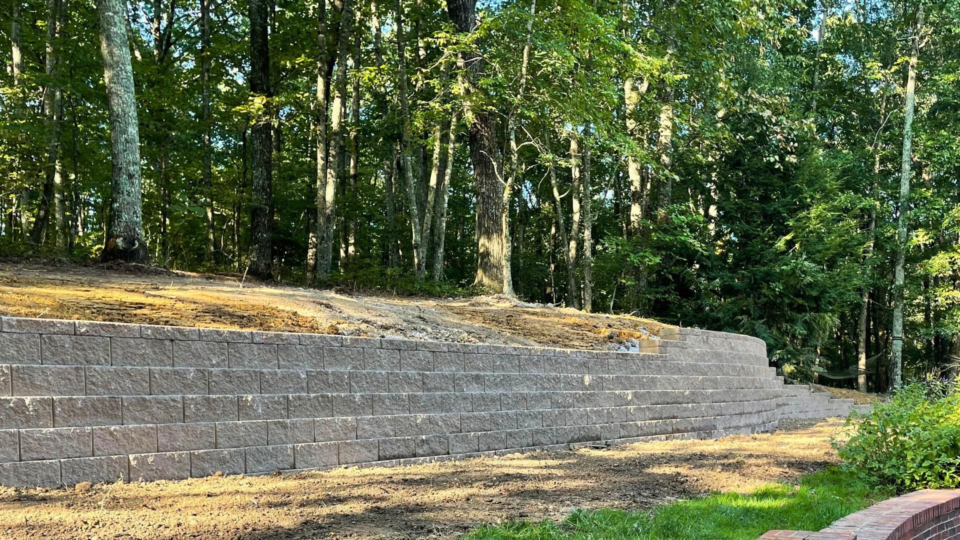 A tall, straight, gray stone retaining wall stands in a wooded yard with freshly leveled dirt in front and behind it.