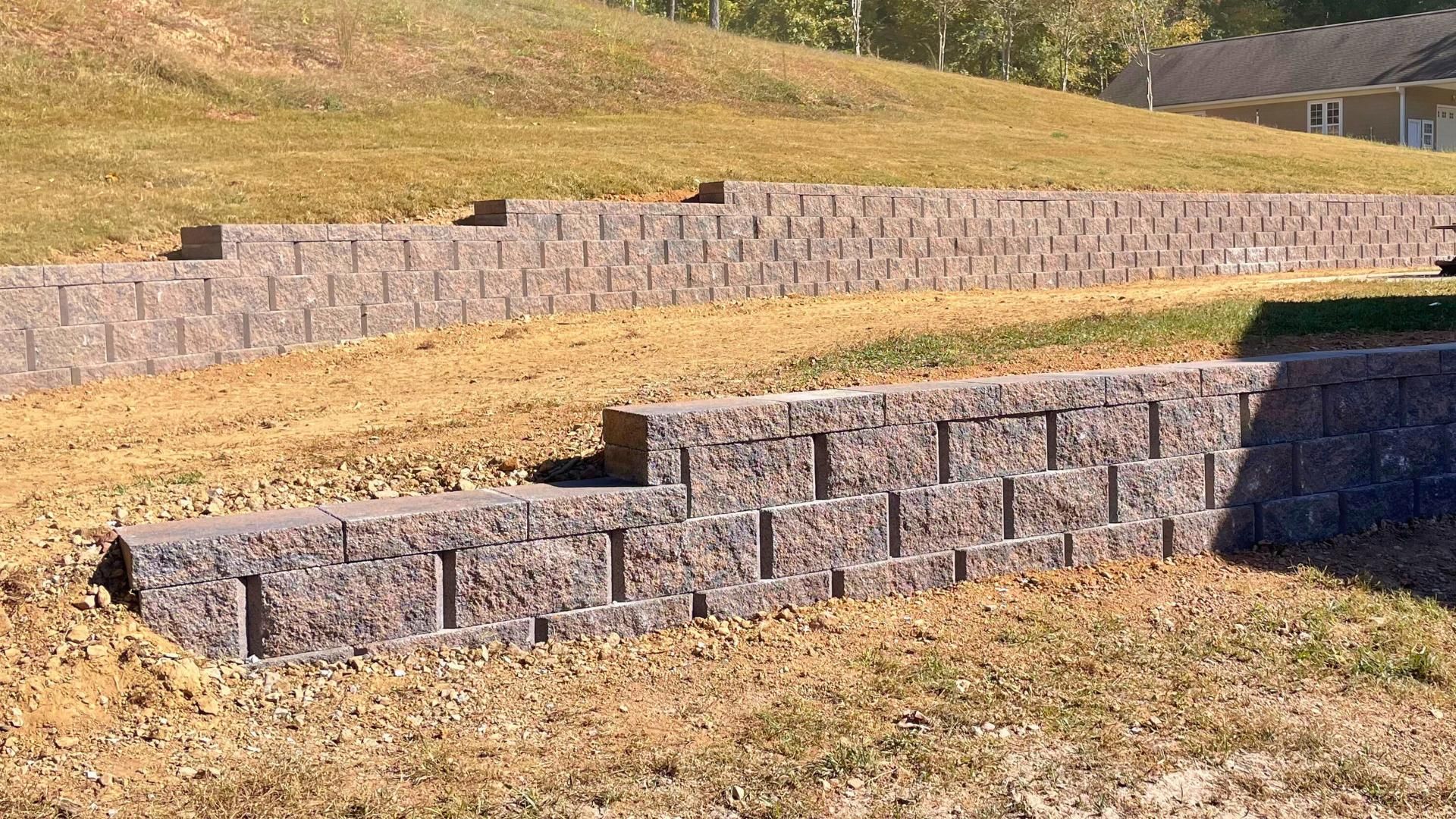 A tiered retaining wall made of textured gray blocks sits on a grassy slope with a house in the background.
