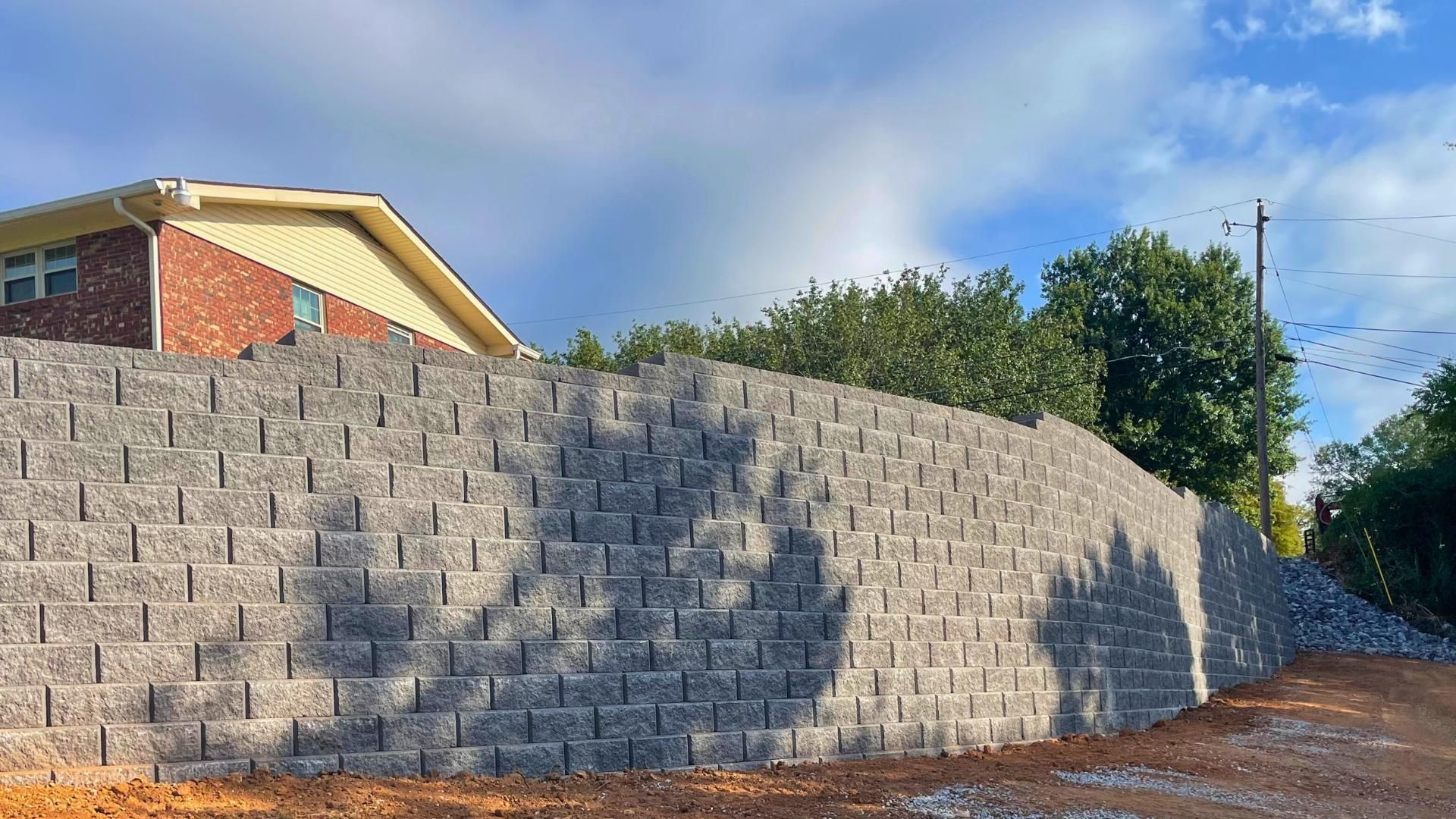 A tall, gray tiered retaining wall made of interlocking concrete blocks sits below a house and trees under a cloudy sky.
