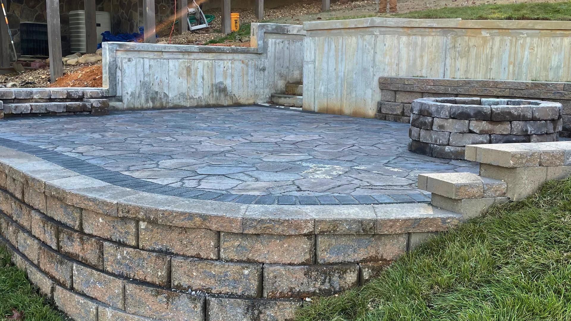 A stone patio with a built-in fire pit, surrounded by beige concrete retaining walls in a grassy yard.