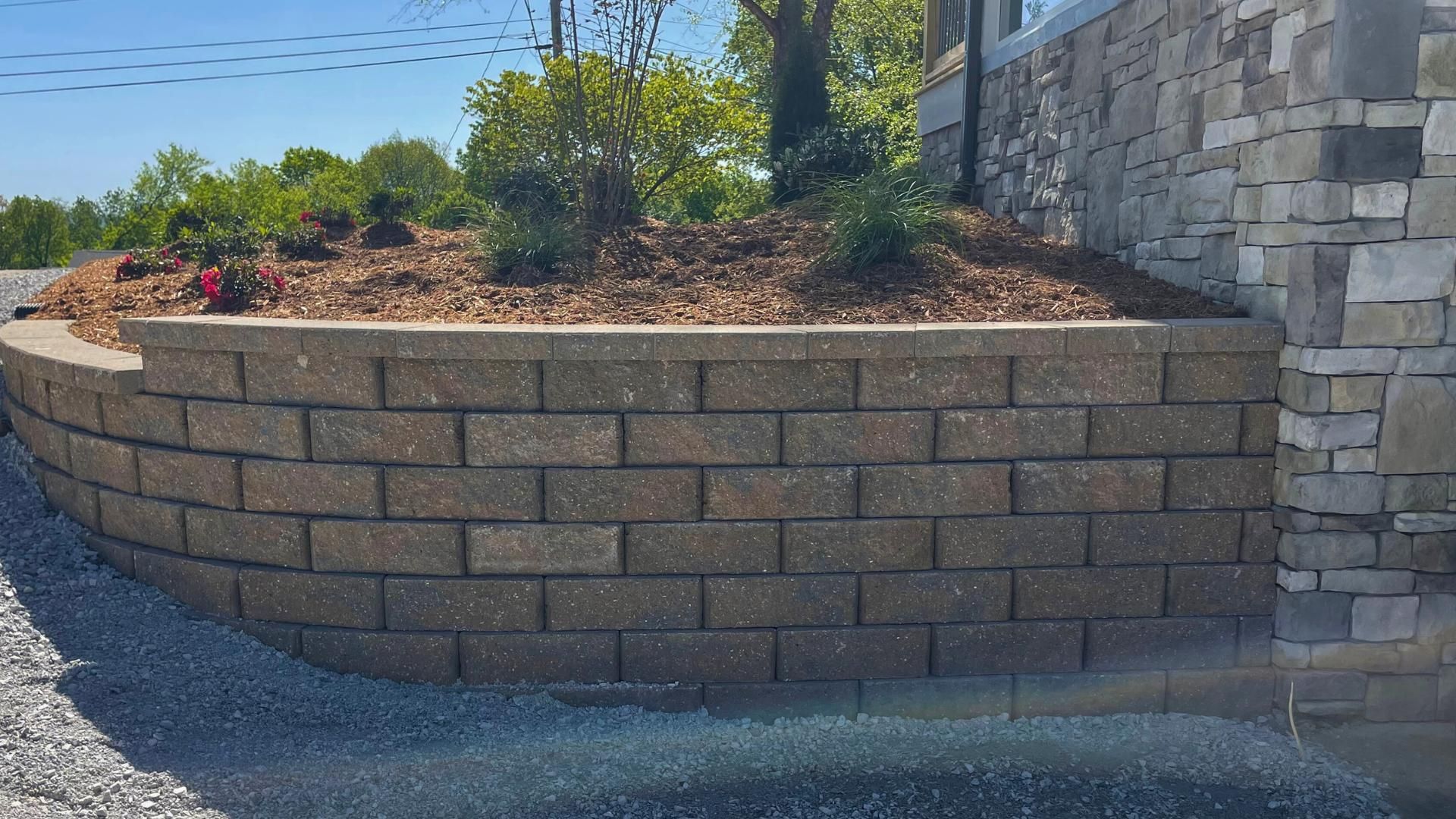 A curved retaining wall made of textured, earth-toned concrete blocks bordering a landscaped garden bed with mulch.
