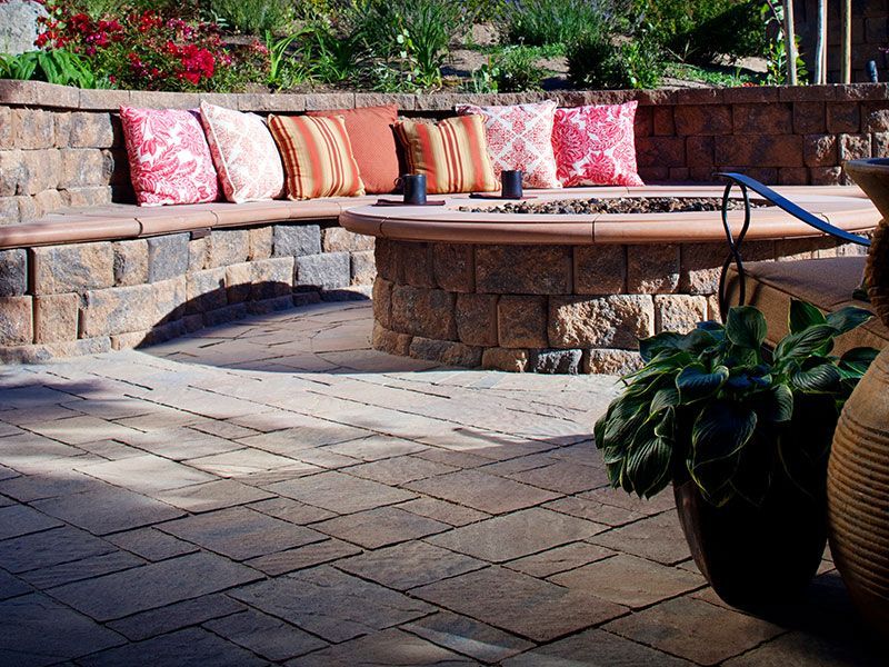 A curved stone seating wall with patterned throw pillows surrounds a circular fire pit on a paved outdoor patio.