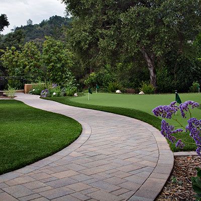 A winding paver walkway curves through a lush green yard with a putting green, trees, and purple flowers.