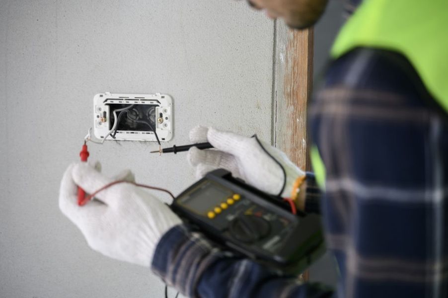 An electrician tests an exposed electrical outlet with a multimeter. They wear gloves and a reflective vest indoors.