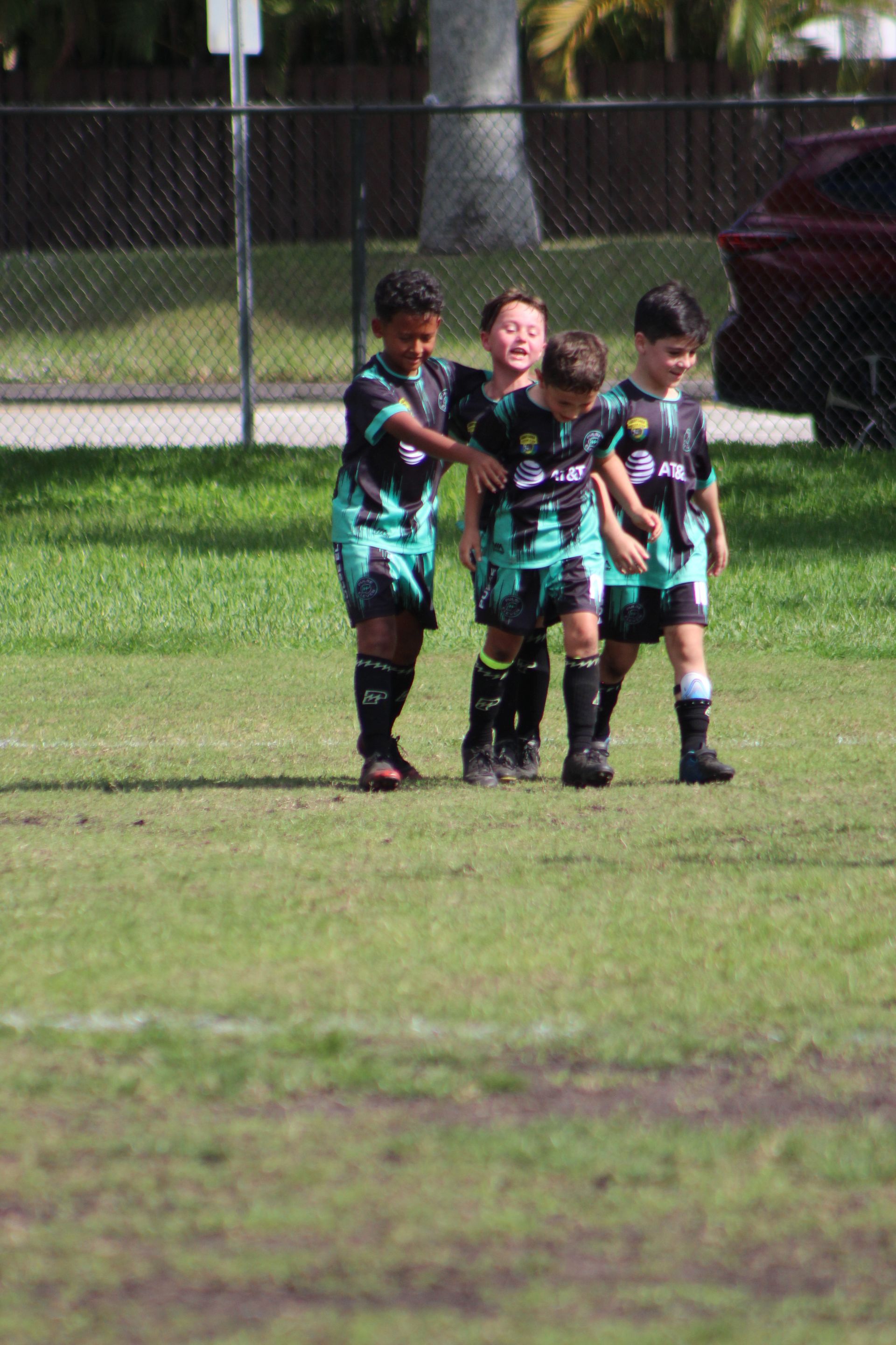 Three children in black and teal sports uniforms standing on a grassy field near a chain-link fence.