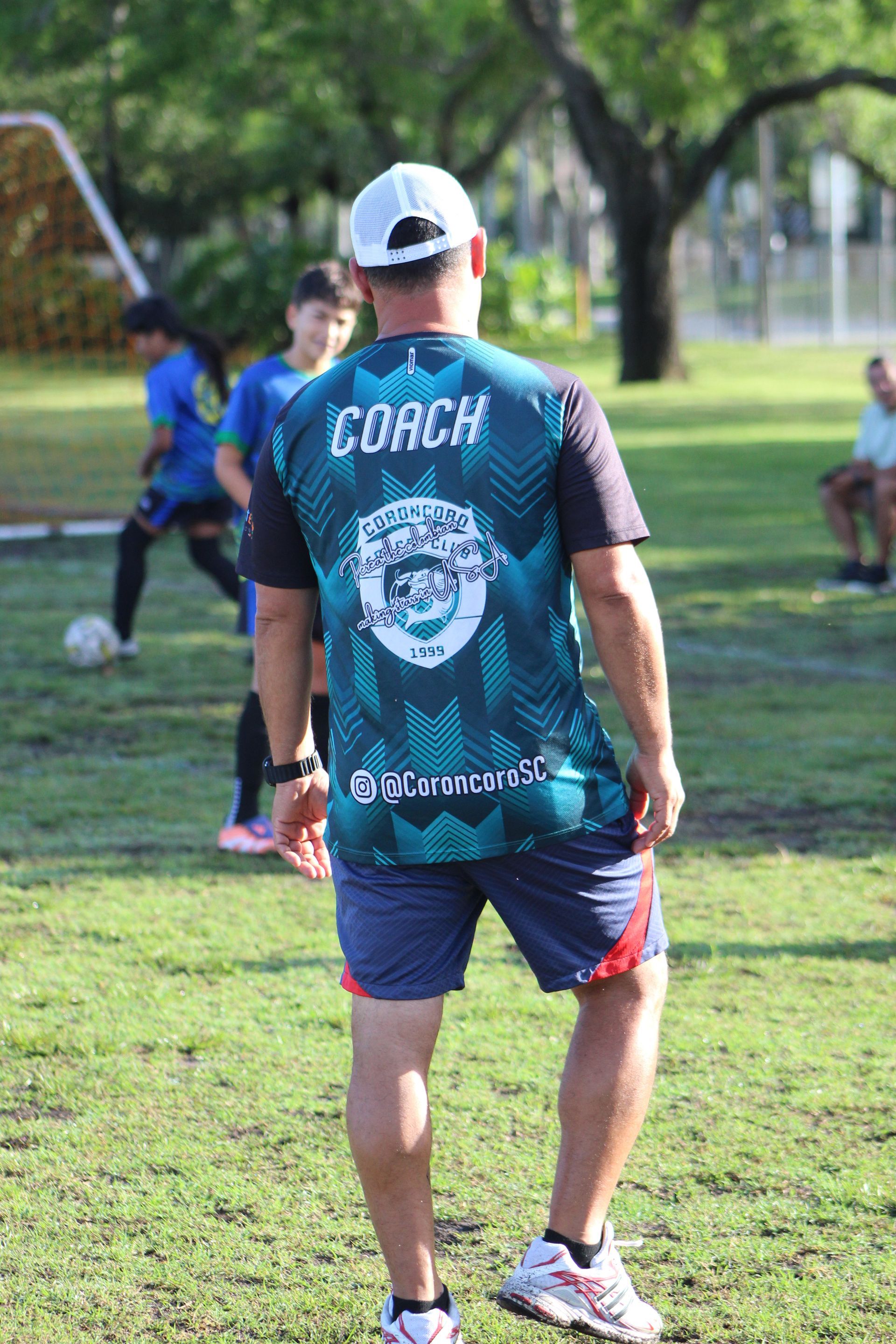 Man in teal sports jersey and white cap standing on a grassy field, viewed from behind.