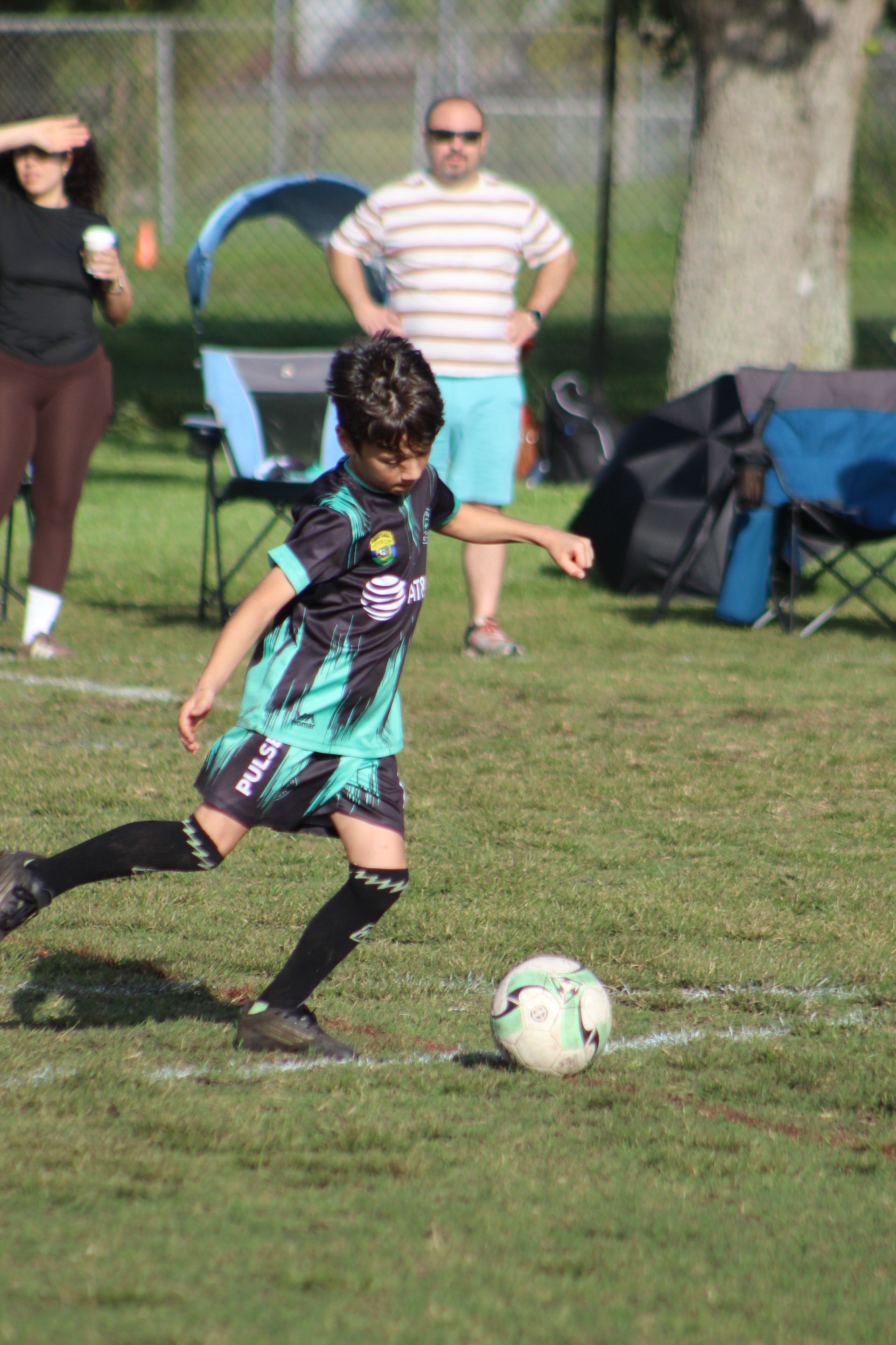 Young soccer player in black and teal uniform kicking a ball on a grassy field during a game