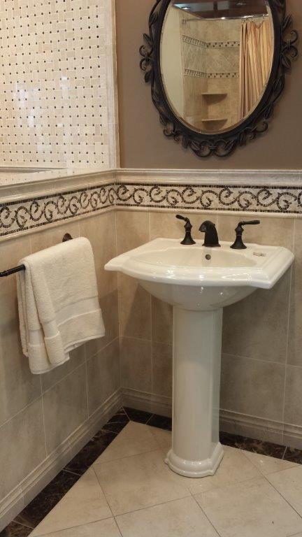 A white pedestal sink with dark faucets sits in a tiled bathroom corner below an oval mirror.