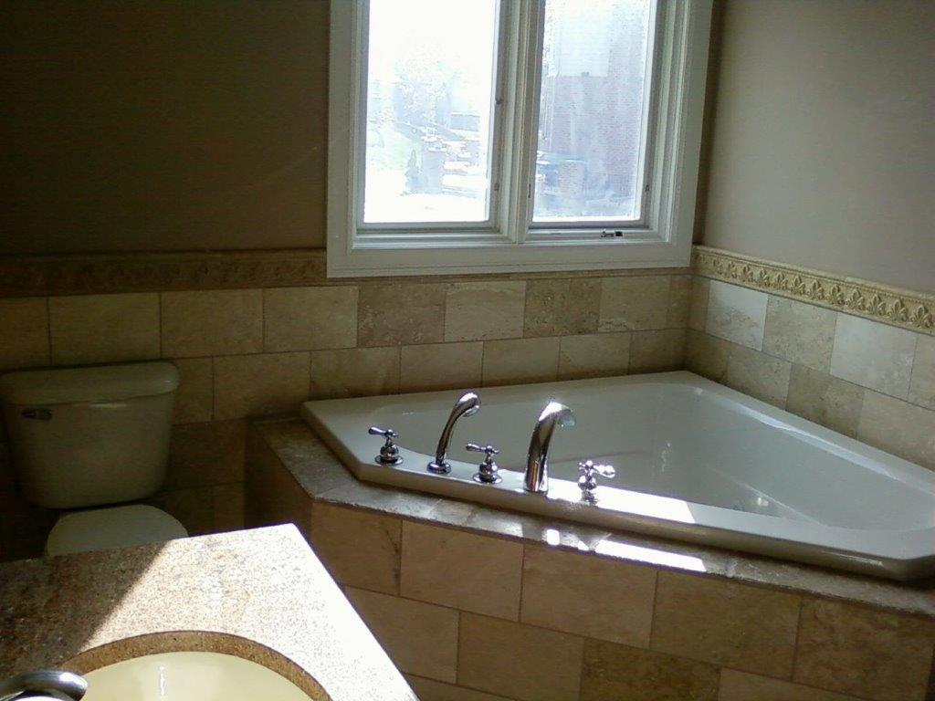A bathroom featuring a corner soaking tub with chrome fixtures, tan stone tile walls, and a window above the tub.