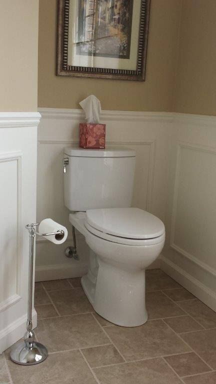 A white toilet in a bathroom with beige walls, white wainscoting, a framed picture, and a chrome toilet paper stand.