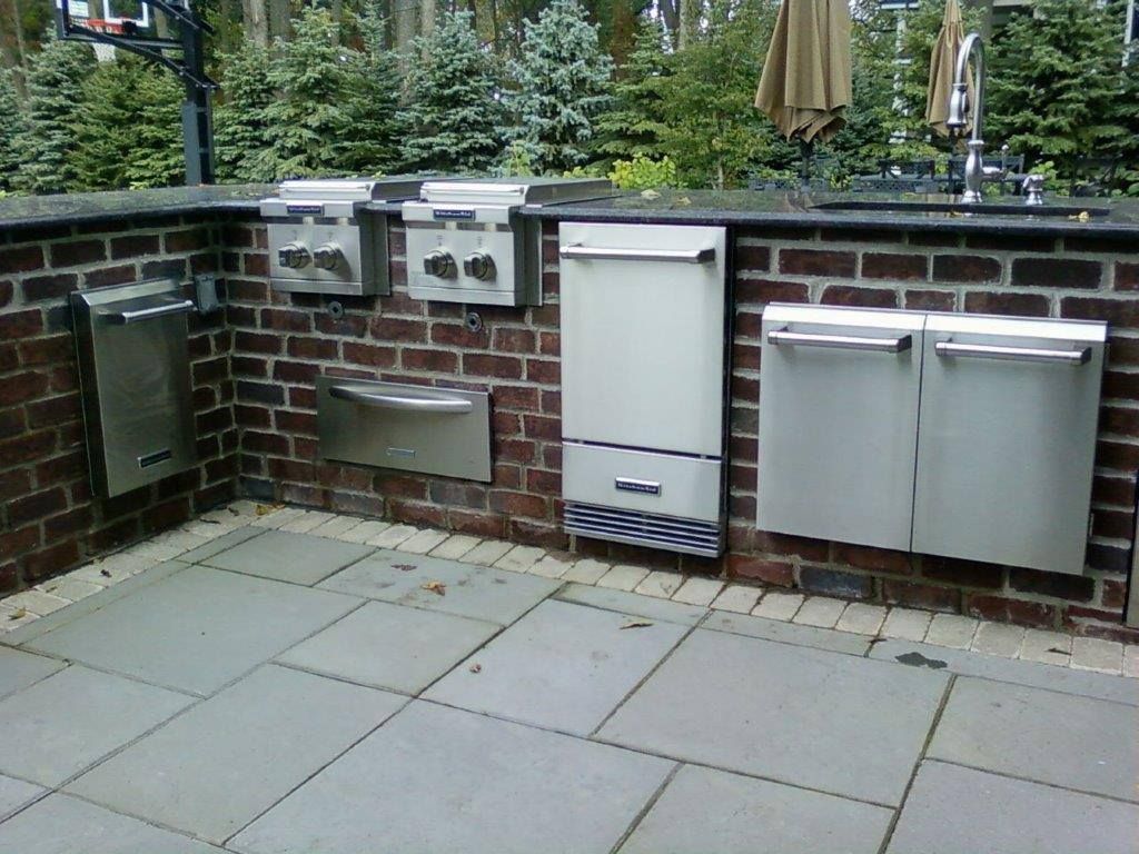An outdoor brick kitchen with stainless steel appliances, including two side burners, a sink, and storage compartments.