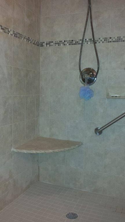 A tiled shower interior featuring a corner seat, a horizontal grab bar, a shower handle, and a decorative mosaic border.