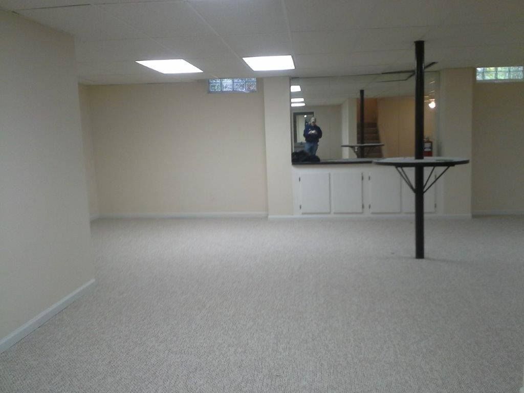An empty basement with beige walls, light-colored carpet, recessed ceiling lights, and a small bar area with a mirror.