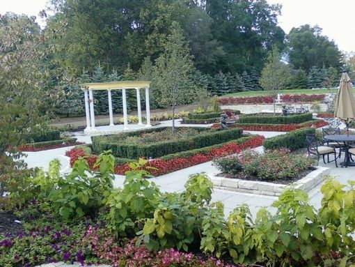 A formal garden with a white classical pergola, neatly trimmed hedges, red flower beds, and a patio dining area.