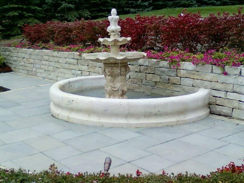 A stone tiered fountain sits in a circular pool on a grey stone patio, set against a low retaining wall and garden beds.