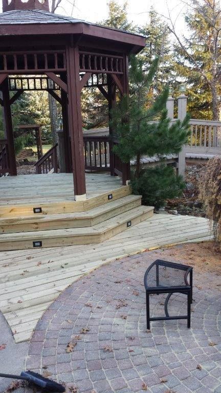 A wooden gazebo with integrated step lights sits on a raised deck next to a curved paver patio with a small metal bench.