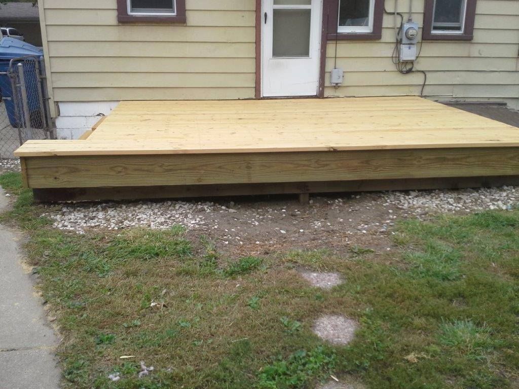 A newly constructed wooden deck sits attached to the rear of a yellow-sided house above a gravel ground area.