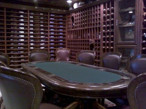 A wooden poker table with a green felt surface sits in a room surrounded by floor-to-ceiling wooden wine racks.