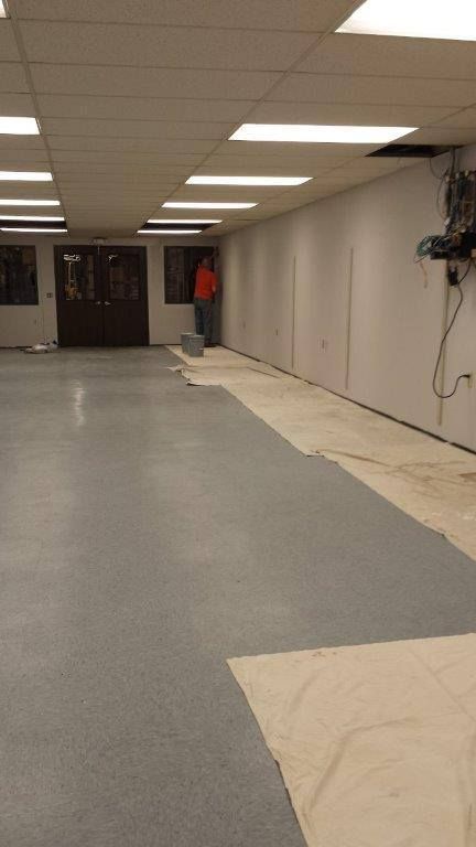 A person in an orange shirt works near a white wall in an office space with grey flooring and drop ceiling tiles.