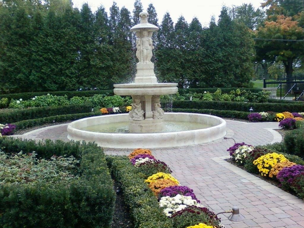 A tiered stone fountain centered in a garden with manicured hedges and rows of colorful flowers along a brick path.