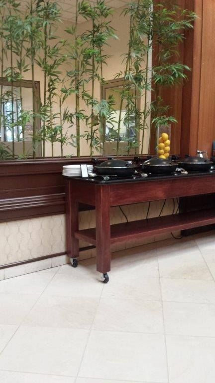 A wooden buffet table with three black serving pans and a stack of plates in front of indoor bamboo plants.