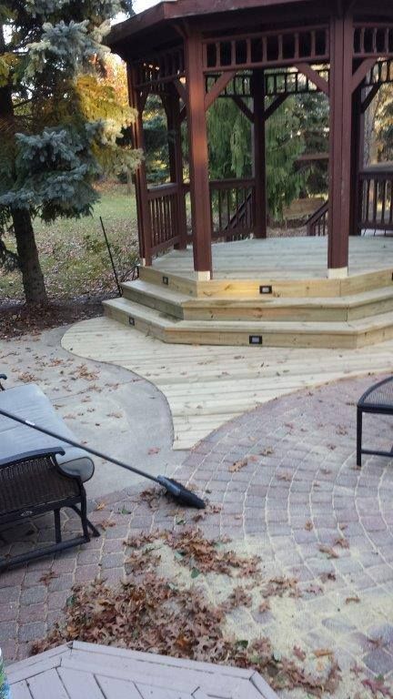 A wooden gazebo with tiered steps sits on a stone patio, with a broom resting nearby amidst scattered autumn leaves.